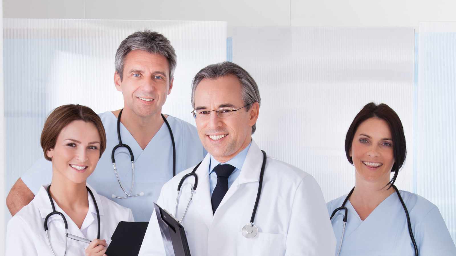 Dwóch lekarzy i dwie lekarki w białych fartuchach lekarskich ze stetoskopami, stojących na tle gabinetu lekarskiego, i patrzących w obiektyw.