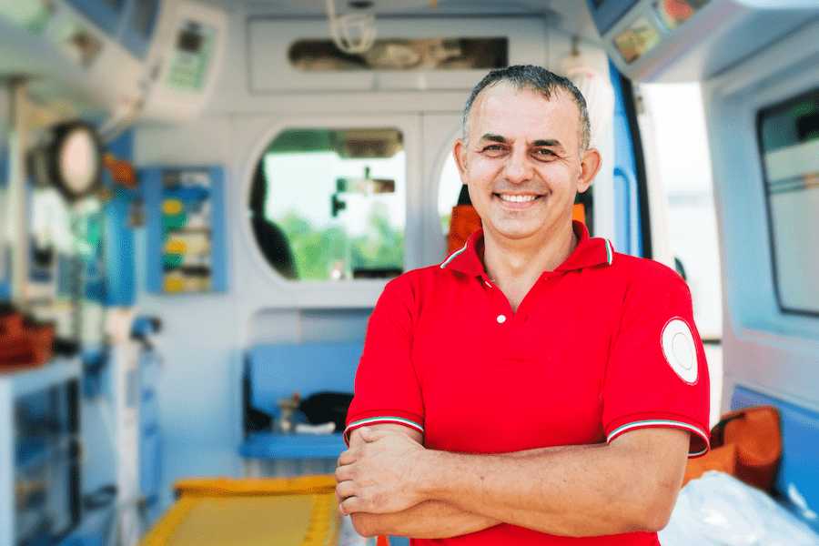 Uśmiechnięty ratownik medyczny, na tle ambulansu medycznego, ubrany w czerwony strój ratownika medycznego, w czasie przerwy od pracy.